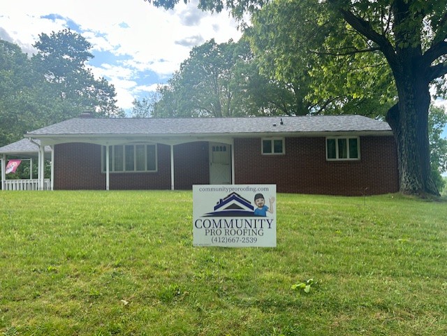 A red brick single-story house with a gray roof stands on a grassy lawn. A large tree provides shade on the right. In the foreground, a sign reads "Community Pro Roofing" with contact information. The sky is partly cloudy.