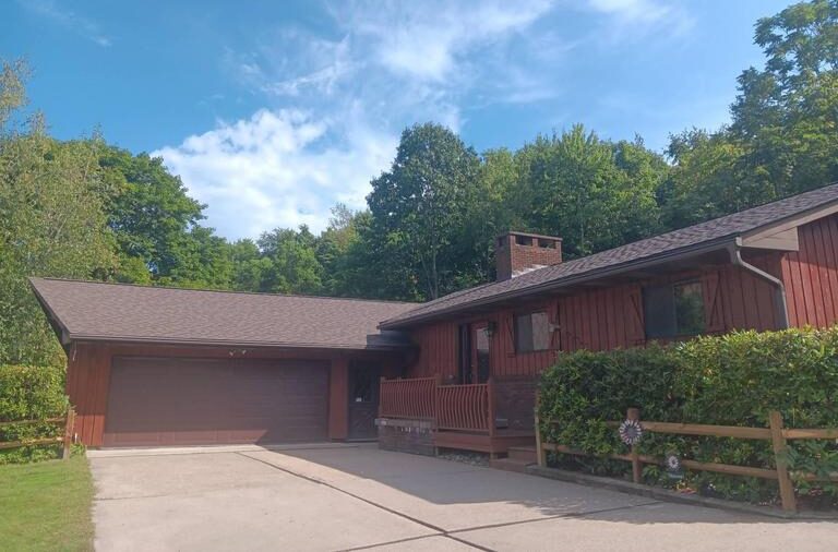 A wooden house with a brown roof and a large garage is surrounded by lush green trees. The sky is partly cloudy, and there is a concrete driveway leading up to the garage. A wooden fence and hedges line the property.
