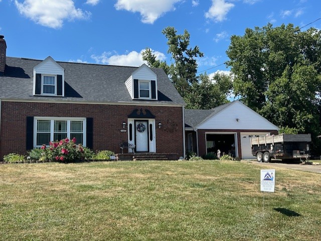 A brick house with a dark roof and two dormer windows. A garage is attached with a trailer parked outside. The front yard is grassy with some flowering shrubs. A sign is placed on the lawn, and trees are in the background under a blue sky.