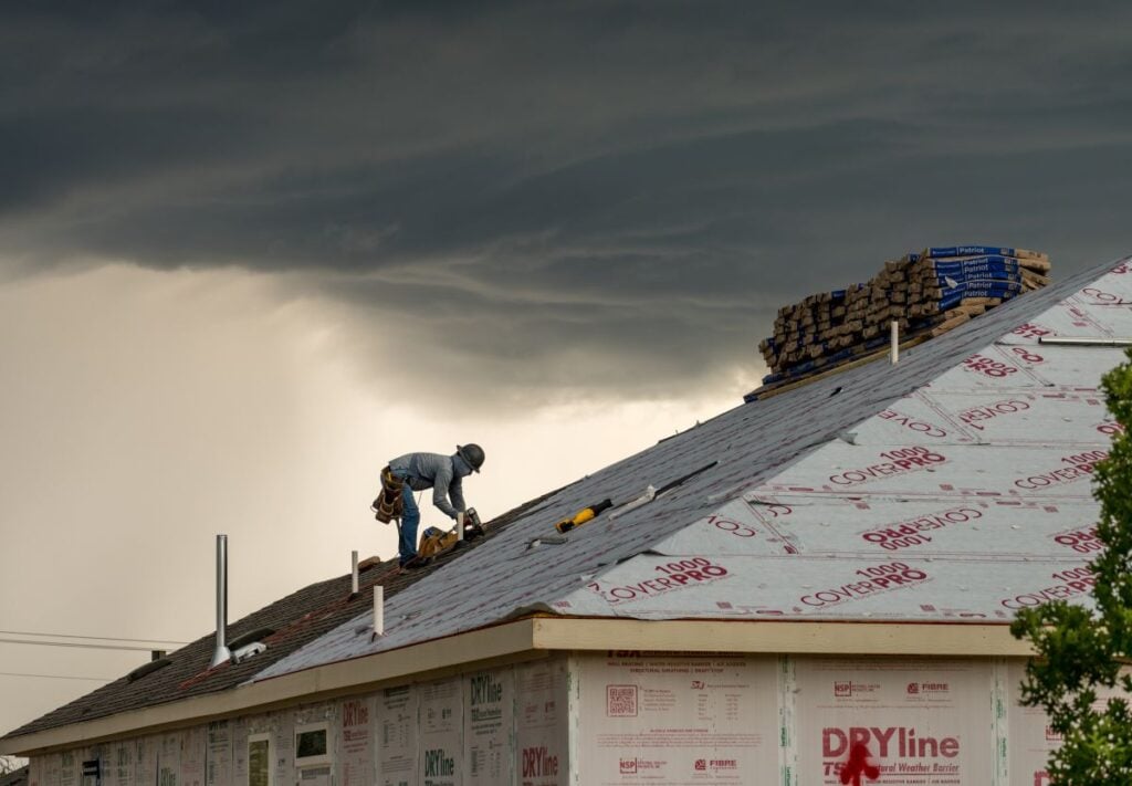 how long does a roof last worker installs roofing material on a house under dark, stormy clouds. Roofing supplies and tools are scattered on the roof, and the house is partially covered in protective wrap.
