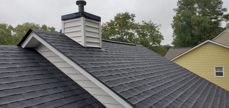 how long does a roof last close-up view of a house roof with dark shingles, a white chimney, and trees and neighboring houses in the background on an overcast day.