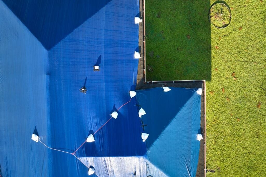 how to tarp a roof Aerial view of blue tarps covering rooftops, secured with sandbags. The tarps contrast sharply with a bright green lawn on the right side of the image. A person and some equipment are visible on the tarps.