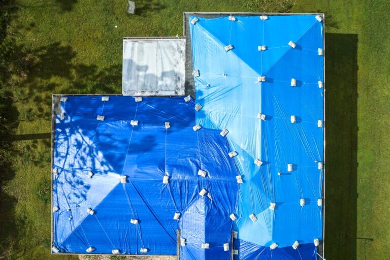 how to tarp a roof Aerial view of a building with its roof covered in large blue tarps held down by sandbags, surrounded by green grass and trees.