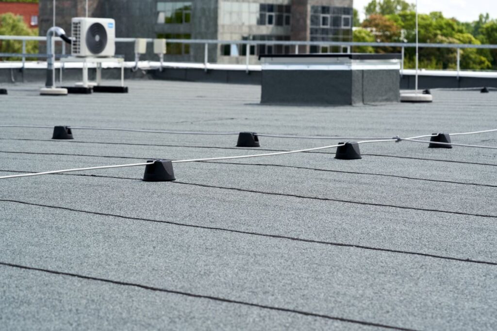 flat roof replacement cost Flat rooftop with a textured gray surface, black safety stanchions connected by a cable, and HVAC equipment in the background, with buildings and trees visible beyond the roof edge.