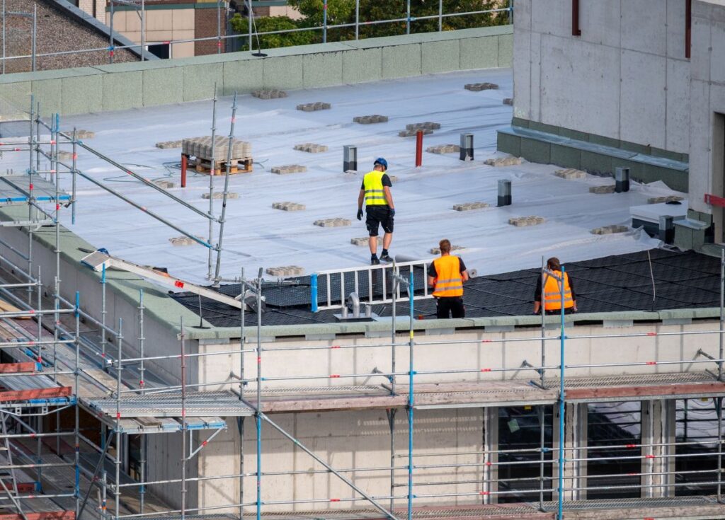 flat roof replacement cost Three construction workers in safety vests and helmets stand on a rooftop under construction, surrounded by scaffolding and building materials. The roof is partially covered with white material.