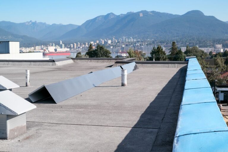 flat roof replacement cost A flat rooftop with ventilation structures overlooks a cityscape and harbor, with forested mountains in the background under a clear blue sky.