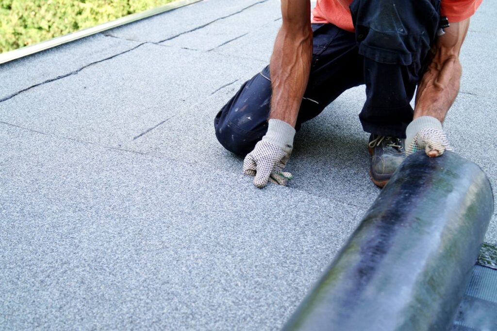 flat roof replacement cost worker wearing gloves kneels on a flat roof, unrolling roofing material during installation or repair. Only the person's arms, hands, and legs are visible. The surface appears gray and textured.