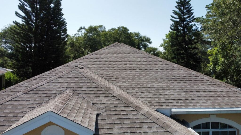 how long does a roof last close-up view of a house roof with brown asphalt shingles, surrounded by tall pine trees and green foliage under a clear blue sky.