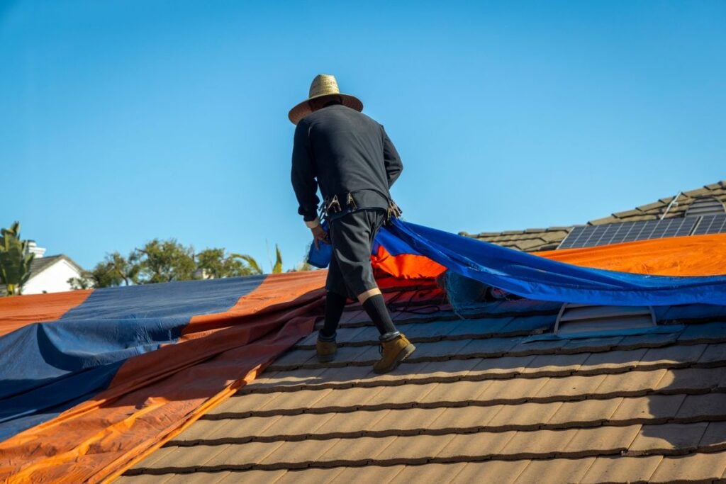 how to tarp a roof A person wearing a wide-brimmed hat and dark clothing secures a large orange and blue tarp on a tiled house roof under a clear blue sky. Some solar panels are visible in the background.