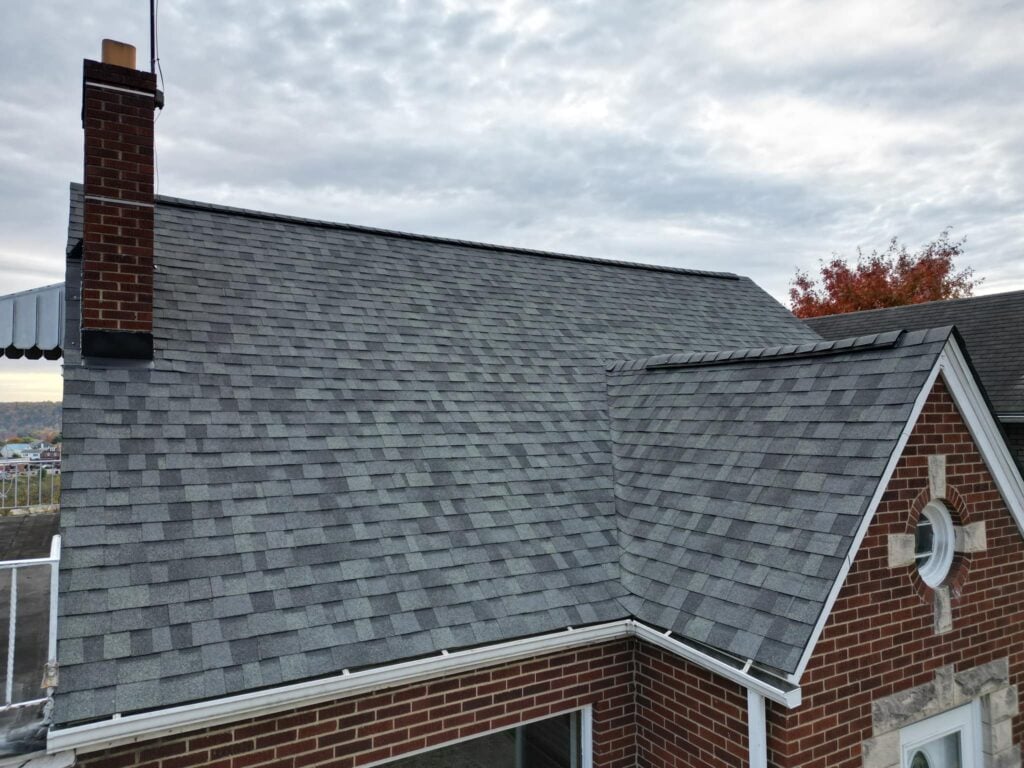 gutter screens A close-up view of a house roof with gray asphalt shingles, a red brick chimney, and part of a brick exterior wall. The sky is cloudy, and some autumn-colored trees are visible in the background.