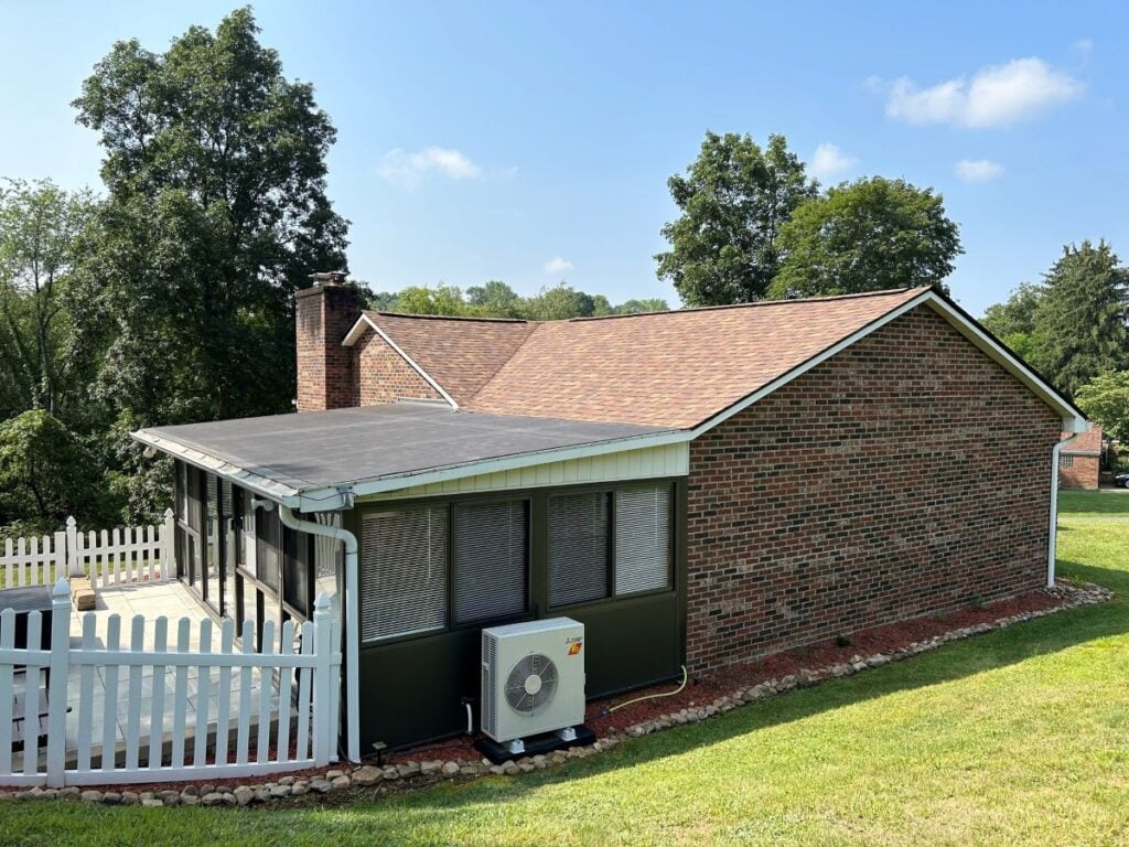 gutter replacement cost A brick house with a screened-in porch, white picket fence, and outdoor air conditioning unit sits on a grassy lawn with trees in the background under a blue sky.