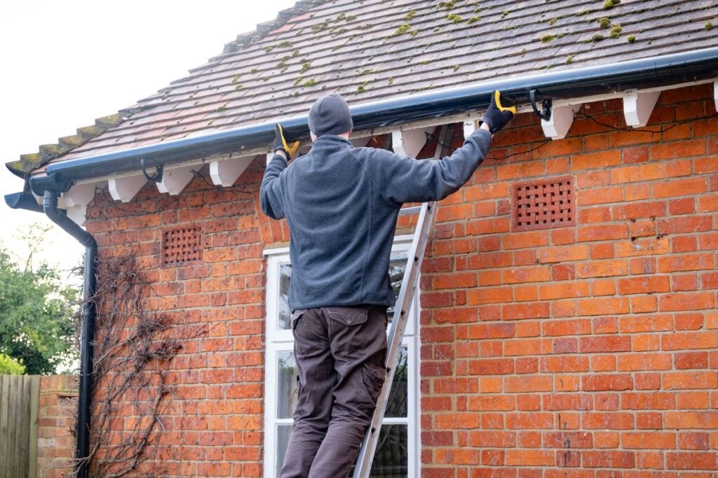gutter replacement cost A person on a ladder cleans or inspects the gutters on a brick house, wearing gloves, a beanie, and a jacket. The roof has some moss, and the weather appears cool.