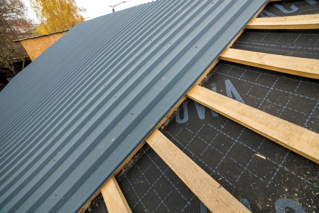 roof underlayment Close-up of a sloped roof under construction with wooden battens, black waterproof membrane, and partially installed gray corrugated metal roofing sheets. Trees and a building are visible in the background.