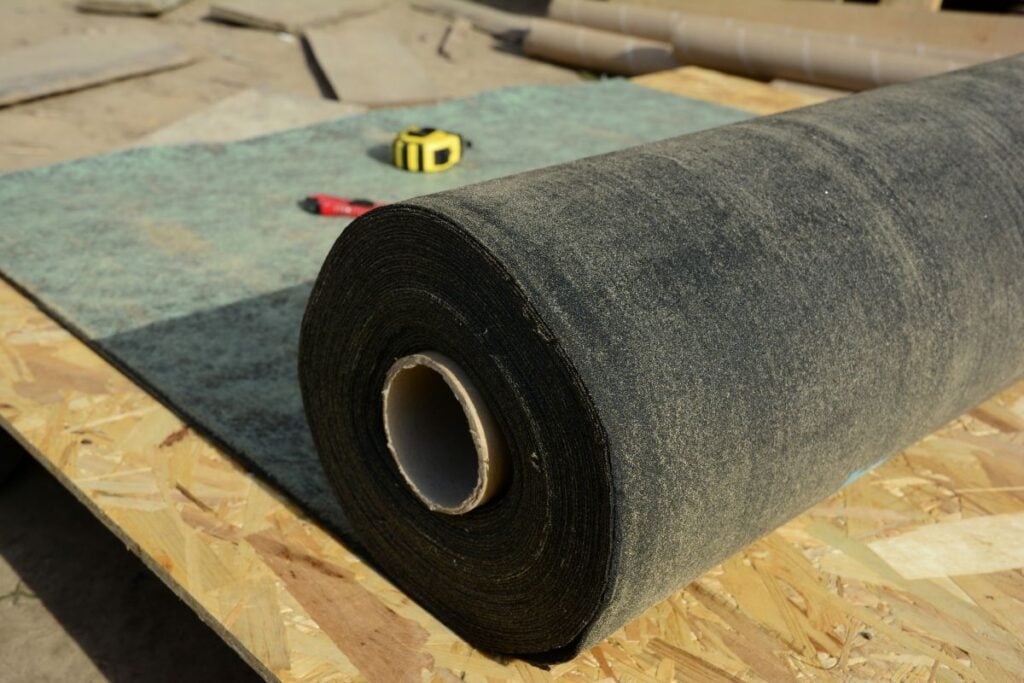 roof underlayment A large roll of roofing felt lies on a sheet of oriented strand board, with a utility knife and a yellow measuring tape in the background.