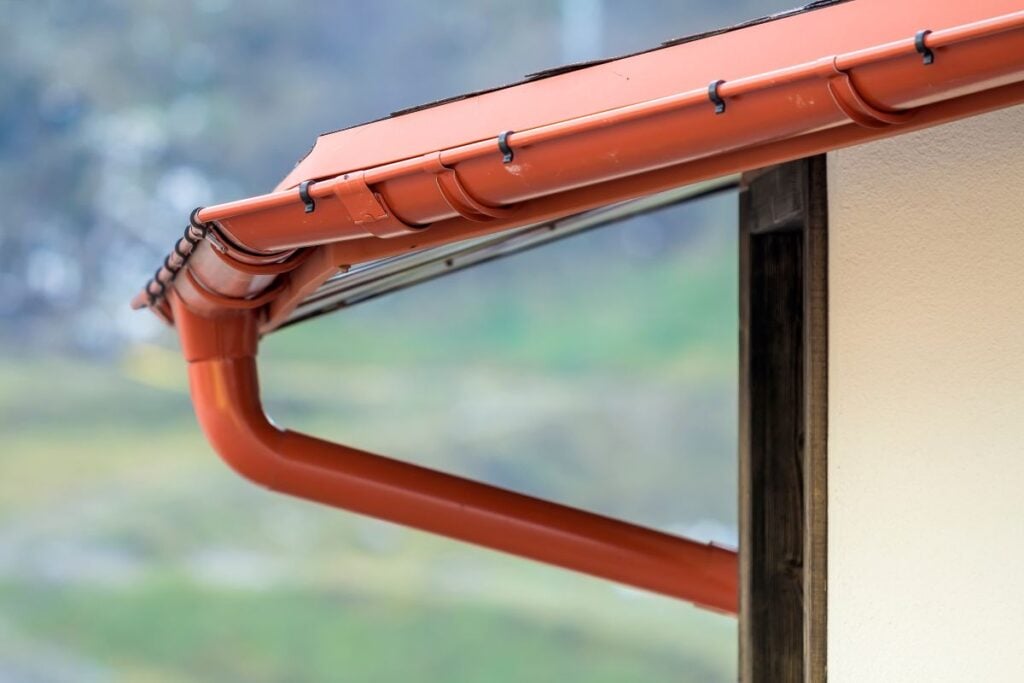 gutter replacement cost Close-up of a house's roof corner showing a clean, red rain gutter and downspout attached to the edge, with a blurred outdoor background of grass and trees.