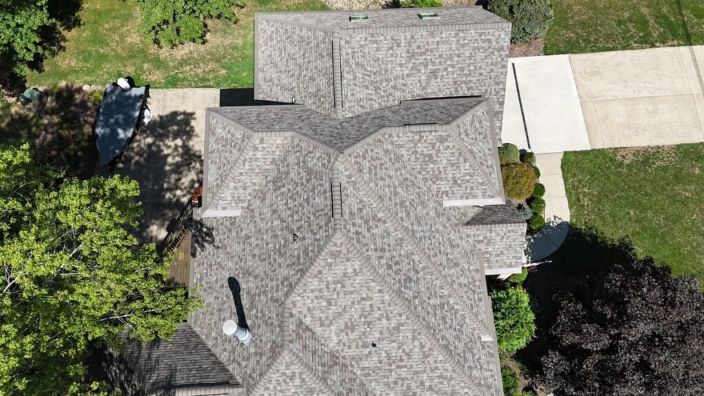 Aerial view of a house roof with complex, intersecting gables and gray shingles, surrounded by trees, lawn, a driveway, and a parked white car visible near the top left corner.