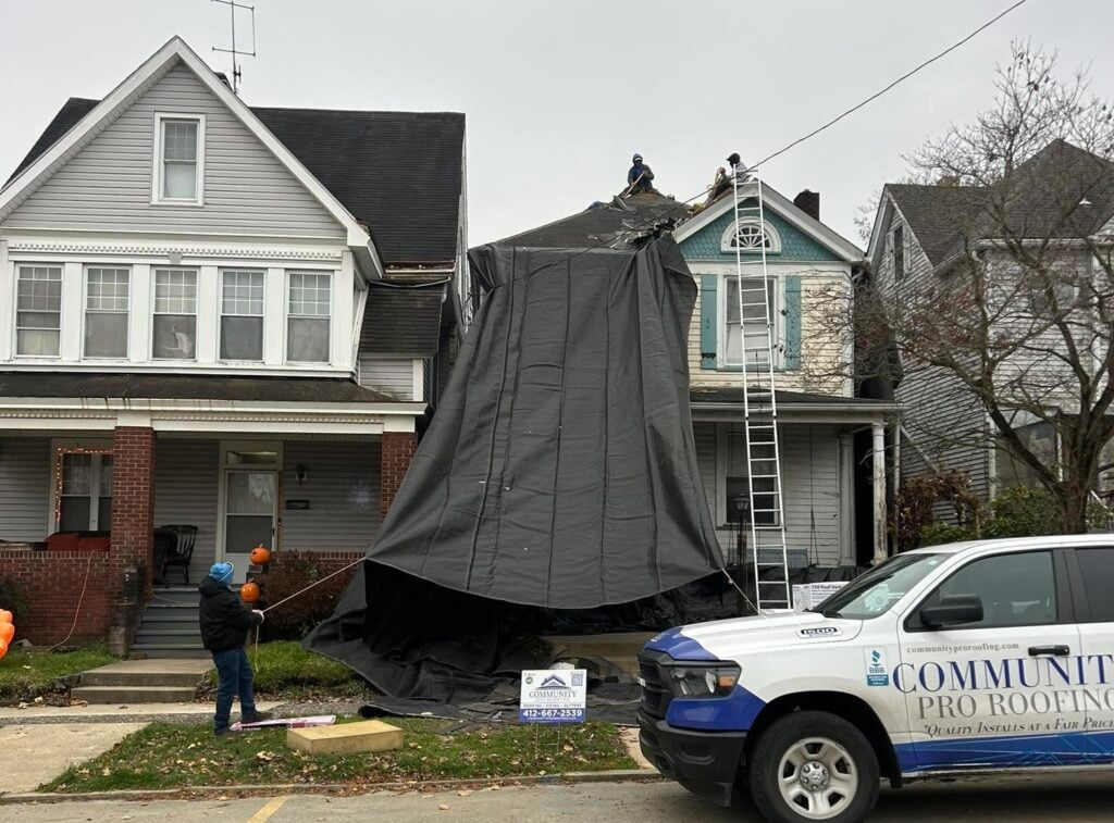 Roofers work on a house with a large black tarp covering its facade, while two people stabilize the tarp below. A Community Pro Roofing truck is parked in front of the house.