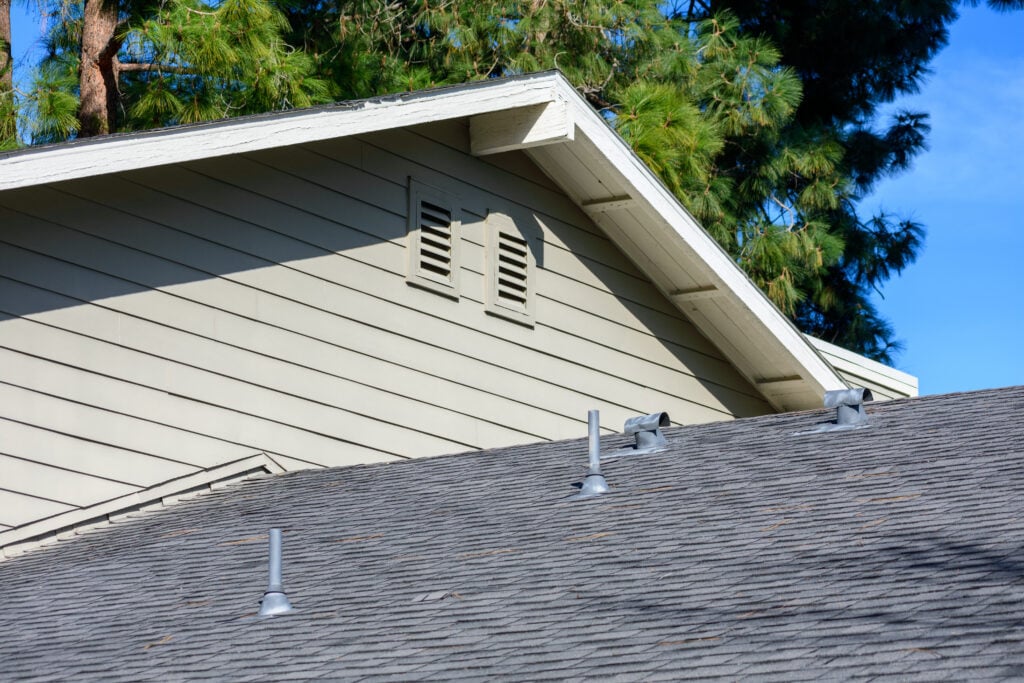 A house roof with gray shingles and several vent pipes, with part of the house’s upper siding and attic vent visible. Green pine trees and blue sky are in the background.