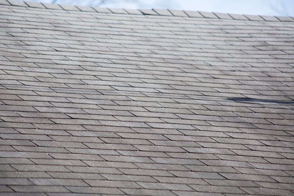 Close-up view of a roof covered with gray asphalt shingles, arranged in neat, slightly overlapping rows. The image captures the texture and pattern of the shingles under daylight.