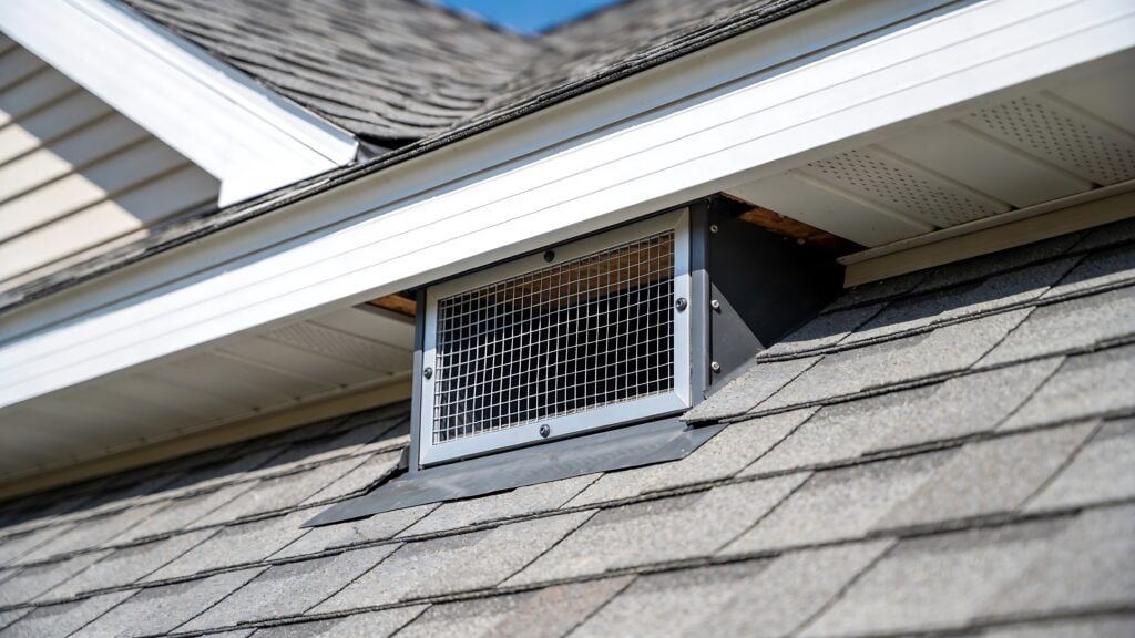 A metal roof vent with a protective wire mesh cover is installed on a shingled roof near the gutter and white soffit under a blue sky.