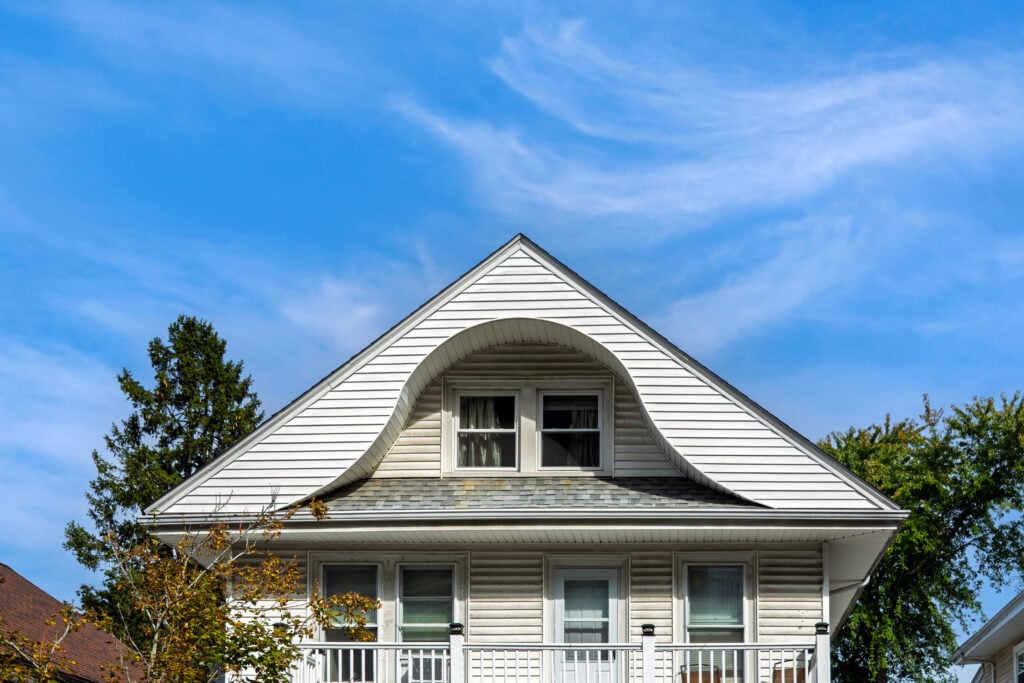 roof types The upper portion of a white house with a unique curved dormer above two windows, under a blue sky with wispy clouds. Trees are visible around the house.
