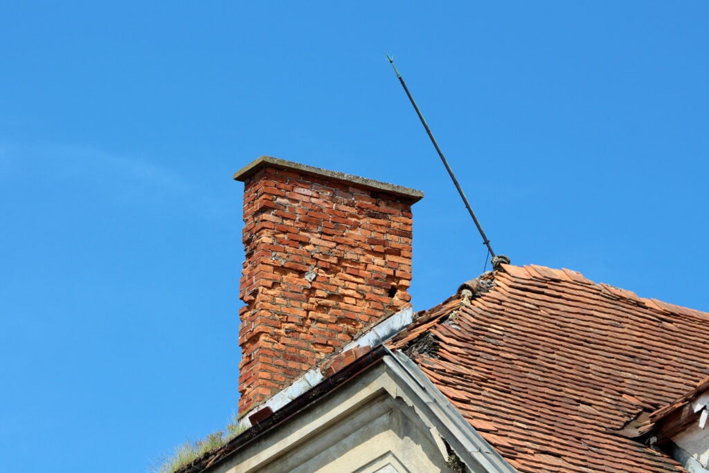 A red brick chimney rises from a sloped, weathered roof with reddish tiles, set against a clear blue sky. A thin metal rod extends upward near the chimney.