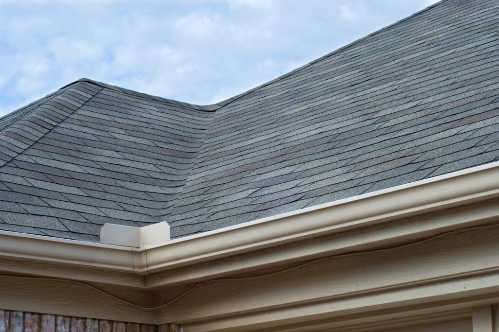 seamless gutter installation Close-up of a house roof with gray asphalt shingles and beige gutters, set against a partly cloudy sky. The shingles are neatly arranged, and part of a brick wall is visible at the bottom left.