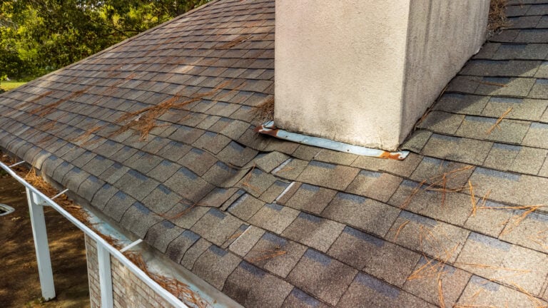 chimney leak repair A close-up of a roof with gray shingles, visible warping near a chimney, and scattered pine needles, indicating possible water damage or structural issues.