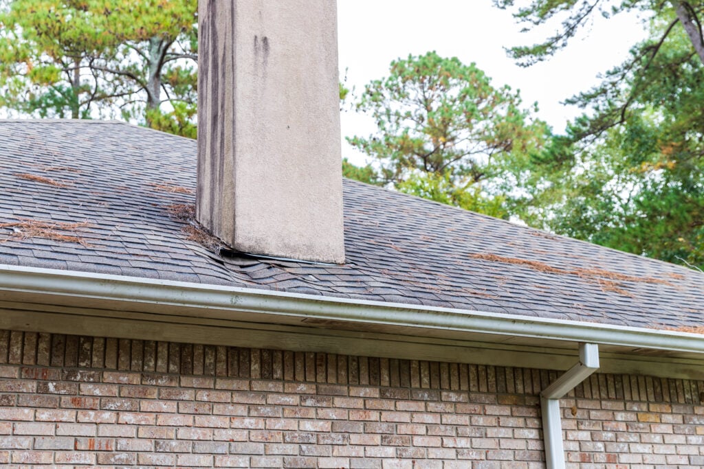 chimney leak repair Chimney with water damage and warped shingles on a house roof, surrounded by pine trees. The brick siding and gutter are visible under the roofline.