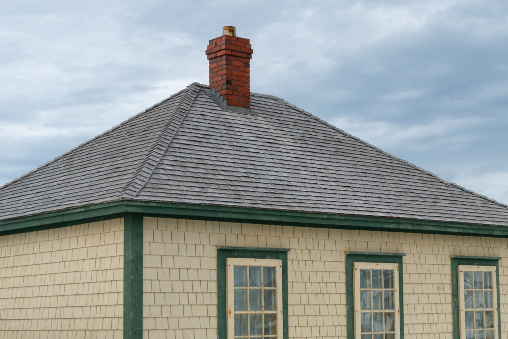 A beige house with green trim, a shingled hip roof, and a red brick chimney is shown against a cloudy sky.