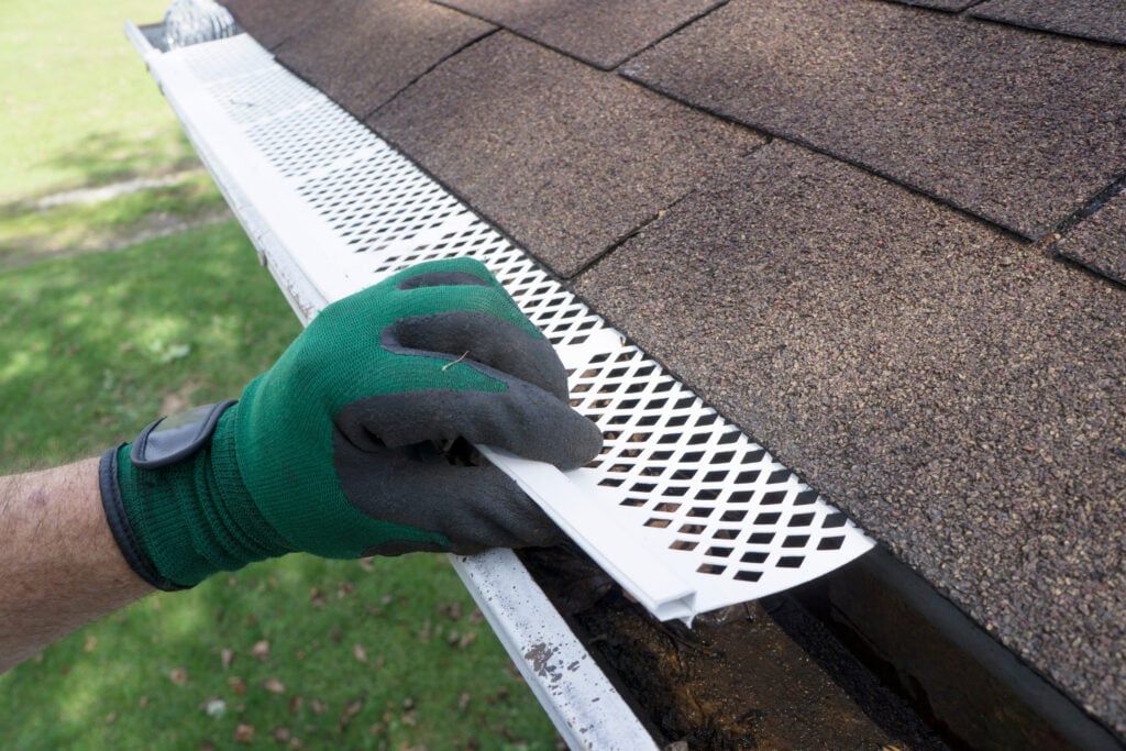 seamless gutter installation A person wearing a green glove installs a white plastic gutter guard on the edge of a shingled roof as part of a seamless gutter installation to prevent debris from clogging the gutter.