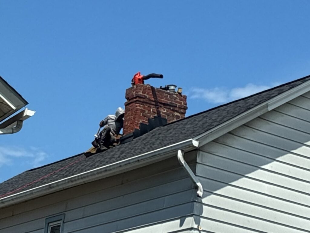 chimney leak repair Two people work on the roof of a house near a red brick chimney under a clear blue sky; one person appears to be using tools while the other holds equipment.