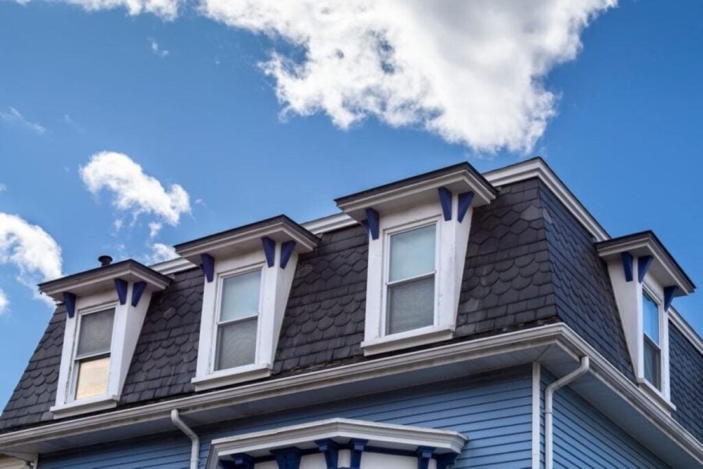 roof types Top floor of a blue house with three dormer windows and decorative trim, featuring dark shingle siding, under a bright blue sky with scattered clouds.