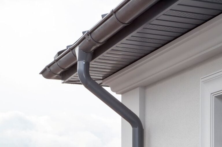 Close-up of a modern house’s roofline featuring a dark gutter and downspout system attached to clean, light-colored eaves and siding, set against a cloudy sky.