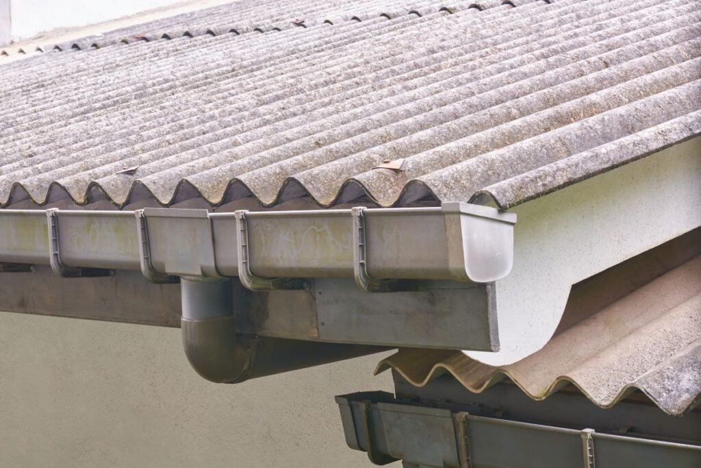seamless gutter installation Close-up of a corrugated asbestos cement roof with a metal gutter system attached to the edge, showing downspout and part of the building’s white wall.