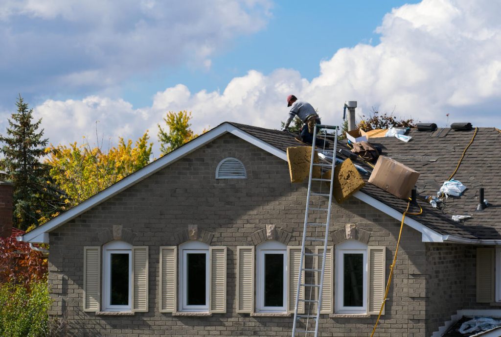 A roofer performs an emergency roof repair on the peak of a residential house with a ladder and discarded materials nearby.